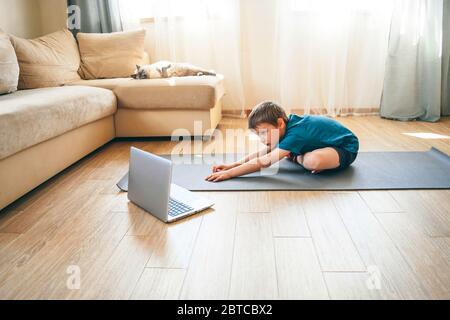 Der Junge macht körperliche Übungen, sieht Video-Lektion online vor offenem Laptop auf dem Boden. Körperliche Übungen auf Distanz. Quarantäne zu Hause. Stockfoto