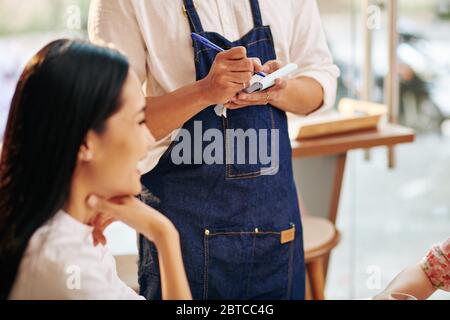 Kellner in Denim-Schürze nimmt Bestellung auf und schreibt sie in Notizbuch auf Stockfoto