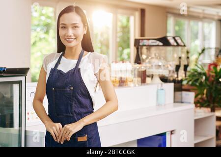 Glückliche erfolgreiche junge Vietnamesin in Denim Schürze stehend in ihrem Coffeeshop oder Café Stockfoto
