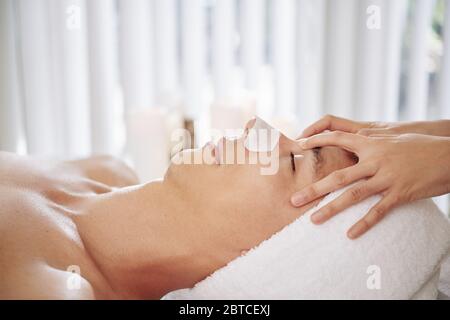 Gemischter junger Mann, der im Spa-Salon Gesichtsbehandlung bekommt und mit einem klebrigen Streifen auf der Nase auf dem Bett liegt Stockfoto