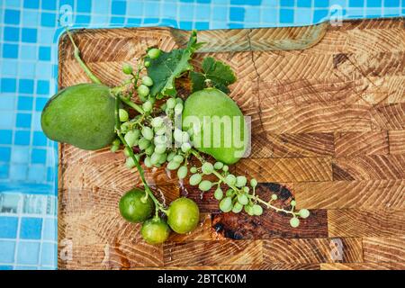 Frische grüne unreife Trauben, Mandarinen und Mangos auf einem Holzhintergrund in blauem Wasser und Kopierraum. Stockfoto