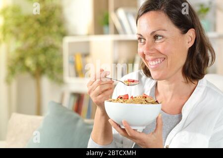 Glückliche Erwachsene Frau mit Müslischale wegschauen beim Frühstück sitzen auf dem Sofa im Wohnzimmer zu Hause Stockfoto