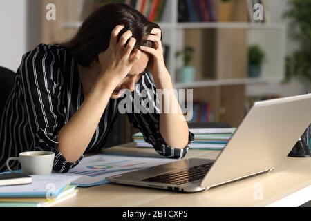 Traurige Unternehmerin mit Laptop, die sich beschwert, nachts zu Hause auf einem Schreibtisch zu sitzen Stockfoto