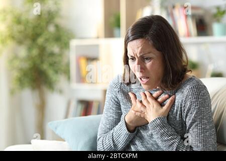 Frau mittleren Alters keuchen, die Brust berühren, die zu Hause auf einer Couch sitzt Stockfoto