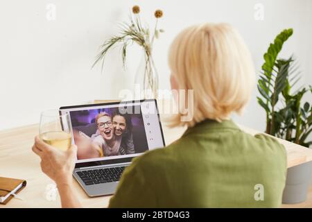 Rückansicht der Frau mit blonden kurzen Haaren, die ein Glas Wein hält und den Urlaub mit ihren Freunden online auf dem Laptop feiert Stockfoto