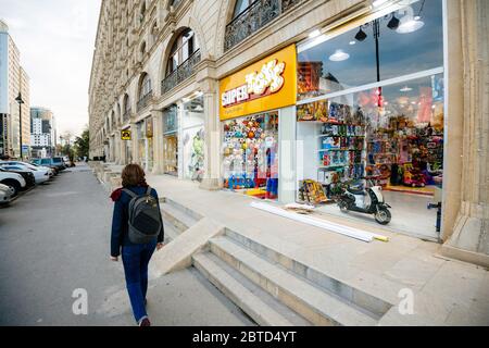 Baku, Aserbaidschan - 2. Mai 2019: Rückansicht der kaukasischen Frau, die auf einer leeren Straße im Zentrum von Baku mit Spielzeugladen in der Nähe und einem großen Apartmentgebäude geht Stockfoto