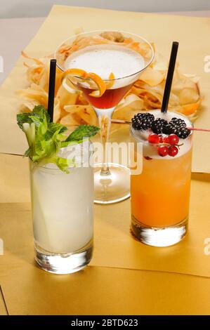 Drei verschiedene alkoholische Cocktails mit frischen Obstgarnieren und einem Teller Kartoffelchips, die auf einer hölzernen Theke in einem hohen Teil gruppiert sind Stockfoto