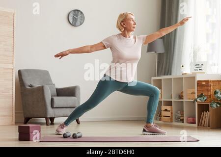 Reife Frau, die auf einer Trainingsmatte steht und Yoga im Wohnzimmer zu Hause macht Stockfoto