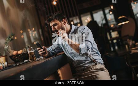 Junger Mann, der Bier trinkt, Zigarette raucht und im Pub Handy benutzt Stockfoto