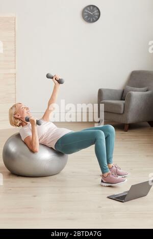 Reife Frau, die beim Online-Training zu Hause mit Hanteln auf dem Ball trainiert Stockfoto