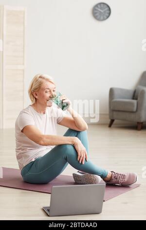 Ältere Frau auf dem Boden auf Trainingsmatte und Trinkwasser nach dem Online-Training zu Hause sitzen Stockfoto