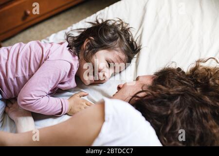 Ein kleines Mädchen, das sich im Bett ihrer Mutter niederlegt, lacht, während sie eine tolle Zeit haben, um zu spielen Stockfoto