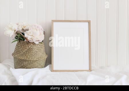 Beige leeres Holzbild Rahmen Modell. Künstlerische Tischstill Life Komposition mit rosa Pfingstrosen Blüten in Strohkorb. Weißer Holzwandhintergrund, Stockfoto