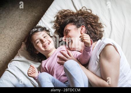 Eine Mutter kitzelt ihre Tochter und lacht laut, während sie auf dem Bett spielt Stockfoto