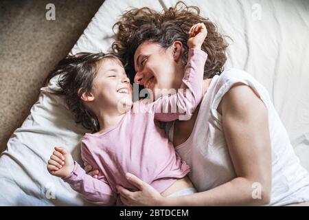 Eine Mutter und ihre Tochter lächeln, während sie sich umschmeicheln und auf einem Bett herumspielen Stockfoto