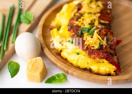 Flauschiges Omelett aus gefaltetem Ei mit Tomate, Paprika, Knoblauch, Frühlingszwiebeln und Cheddar-Käse in einem Holzteller. Stockfoto