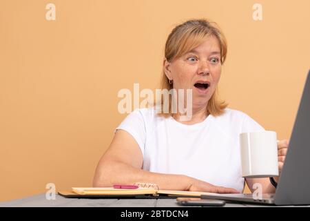 Ältere Frau im mittleren Alter sitzt am Tisch zu Hause mit Computer-Laptop arbeiten, hält Tasse Kaffee Angst und schockiert mit überraschenden Ausdruck Stockfoto