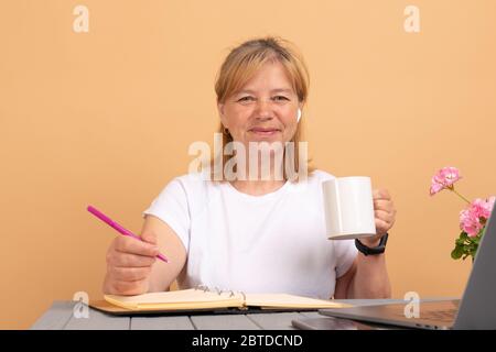 Optimistische Frau in kabellosen Kopfhörern und legeren weißen Kleidern trinken Tasse frischen Kaffee, während sie am Holztisch neben Laptop sitzt und anschaut Stockfoto