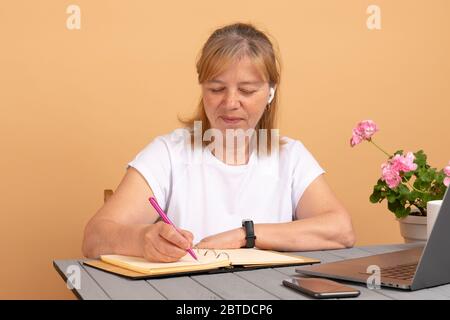 Lächelnde ältere Frau mit Kopfhörern, die mit einem Laptop zu Hause Audiokurse hören, Notizen machen, Frauen mittleren Alters, die Fremdsprachen lernen Stockfoto