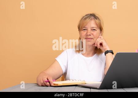 Attraktive positive talentierte reife Schriftstellerin sitzt vor dem Laptop und schreibt ihr neues Buch, Blick auf die Kamera mit freudigem Lächeln Stockfoto