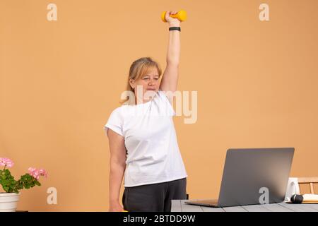 Schöne Frau mit gelben Hanteln in den Händen. Fitness-Training zu Hause Videos online ansehen Arbeiten auf Laptop-Computer - Frau Multitasking Stockfoto
