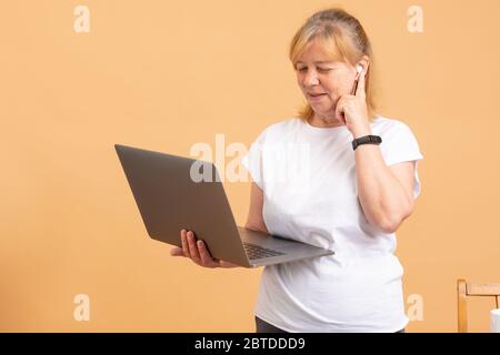 Großmutter im digitalen Zeitalter. Nahaufnahme einer nachdenklichen älteren Frau, die versucht, mit dem Computer fertig zu werden, während sie vor orangefarbenem Hintergrund steht Stockfoto