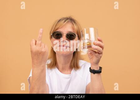Frau mittleren Alters in schwarzer Sonnenbrille hält Glas Whisky in der Hand, zeigt Mittelfinger an der Kamera Stockfoto