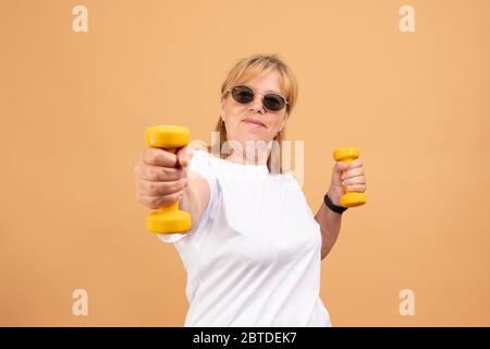 Aufgeregt Senior Fitness Frau in Sonnenbrillen Training mit Kurzhanteln isoliert auf orange Stockfoto