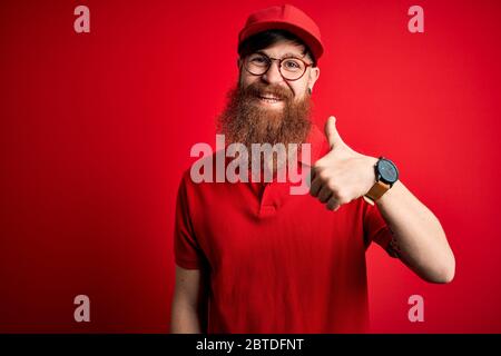 Junge schöne Lieferung Mann trägt Brille und rote Kappe über isolierten Hintergrund tun glücklich Daumen hoch Geste mit der Hand. Suche nach Ausdruck wird genehmigt Stockfoto