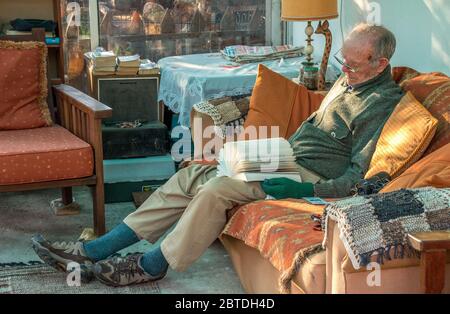 Alberton, Südafrika - alter kaukasischer Mann, der auf einer Couch in der Sonne schläft, mit einem offenen Buch auf seinem Schoß, im horizontalen Format Stockfoto