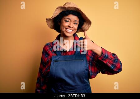 Junge afro amerikanische Afro Farmer Frau mit lockigen Haaren tragen Schürze und Hut tun glücklich Daumen hoch Geste mit der Hand. Genehmigen eines Ausdrucks, der nach einem sucht Stockfoto