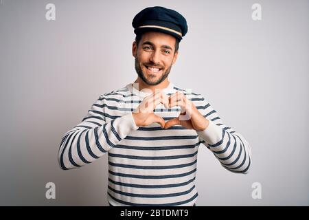 Junge schöne Seemann mit Bart trägt marineblau gestreifte Uniform und Kapitän Hut lächelnd in der Liebe zeigt Herz Symbol und Form mit den Händen. Romantisch Stockfoto