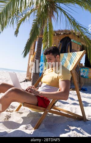 Kaukasischer Mann, der auf einem Liegestuhl sitzt und einen Laptop am Strand benutzt Stockfoto