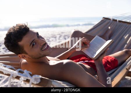 Kaukasischer Mann, der auf einer Hängematte liegt und am Strand ein Buch liest. Stockfoto