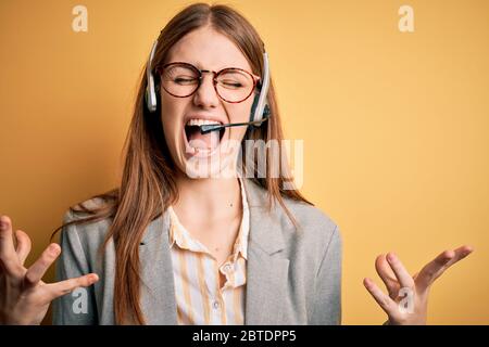 Junge Redhead Call Center Agent Frau überfordert tragen Brille mit Headset verrückt und verrückt schreien und schreien mit aggressivem Ausdruck und Arme Stockfoto