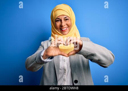 Mittelalter Brünette Geschäftsfrau trägt muslim traditionellen Hijab über blauem Hintergrund lächelnd in Liebe tun Herz Symbol Form mit Händen. Romantisch Stockfoto