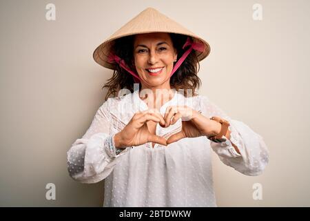 Mittelalter Brünette Frau trägt asiatische traditionelle konische Hut über weißem Hintergrund lächelnd in Liebe tun Herz Symbol Form mit Händen. Romantisches CO Stockfoto