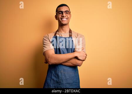 Junge gut aussehende afroamerikanischen Ladenbesitzer Mann trägt Schürze über gelben Hintergrund glückliches Gesicht lächelnd mit verschränkten Armen Blick auf die Kamera. Positi Stockfoto