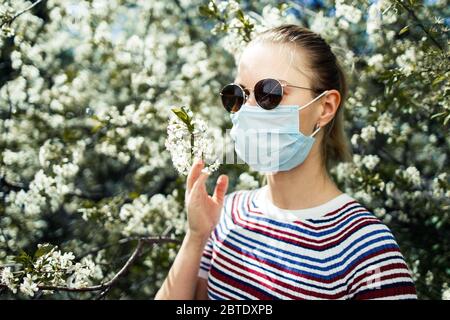 Frau in medizinischer Maske und Sonnenbrille auf dem Hintergrund des blühenden Apfelbaums am Sommertag. Stockfoto