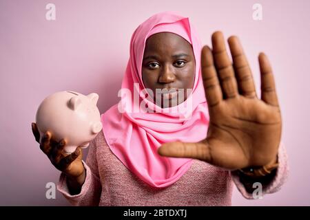 Junge african american plus size Frau trägt muslimischen Hijab mit rosa Sparschwein mit offener Hand tun Stop-Zeichen mit ernsten und zuversichtlich expressi Stockfoto