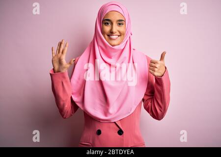 Junge schöne Brünette Geschäftsfrau trägt rosa muslimischen Hijab und Business-Jacke zeigt und zeigt mit den Fingern Nummer sechs beim Lächeln con Stockfoto