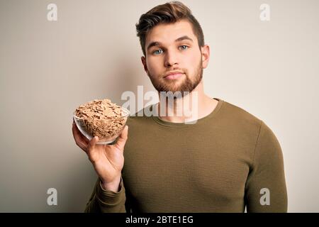 Junge blonde Mann mit Bart und blauen Augen halten Schüssel mit Cornflakes Getreide mit einem selbstbewussten Ausdruck auf intelligente Gesicht denken ernst Stockfoto