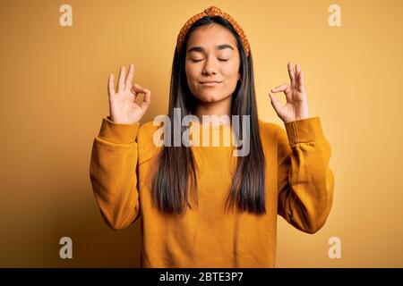 Junge schöne asiatische Frau in lässigen Pullover und Diadem über gelben Hintergrund entspannen und lächeln mit geschlossenen Augen tun Meditation Geste mit Stockfoto