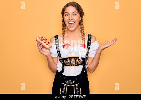 Schöne blonde deutsche Frau mit blauen Augen trägt oktoberfest Kleid essen Hot Dog sehr glücklich und aufgeregt, Sieger Ausdruck feiern Sieg sc Stockfoto