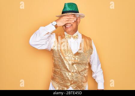 Alter älterer grauhaariger Mann, der brasilianische Karnevalskumsty über gelbem Hintergrund trägt und lächelt und lacht mit Hand auf Gesicht, das Augen für s bedeckt Stockfoto