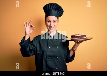 Junge schöne Bäckerin Frau trägt Herd Uniform und Hut halten Tablett mit Kuchen tun ok Zeichen mit Fingern, ausgezeichnetes Symbol Stockfoto
