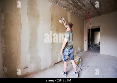 Rückansicht des Kerls, der Handwerkzeug-Spray der Farbe hält und eine Wand tagsüber malt. Männlich ist in Farbe verschmierte Kleidung gekleidet. Haus renoviert. Stockfoto