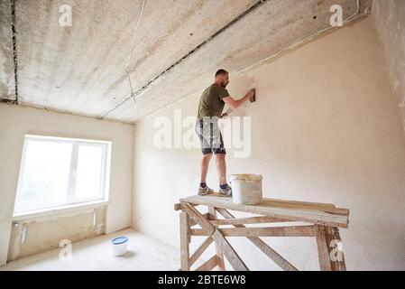 Baumeister Mann steht auf einem Holzständer und arbeitet mit einem Spachtel mit Gips an der Wand gegen ein Fenster. Die Wände innen verputzen. Ein Typ mit Bart im T-Shirt und Jeans ist mit Farbe verschmiert Stockfoto