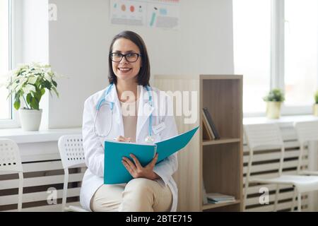 Porträt der jungen Ärztin, die ein offenes Standesbuch hält und lächelnd in der Kamera sitzt, während sie im Empfangsbereich der modernen Klinik sitzt, kopieren Raum Stockfoto