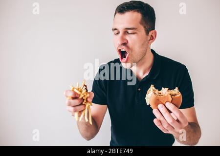 Junger Mann isoliert auf weißem Hintergrund. Hungriger Kerl, der Pommes und Burger in den Händen hält. Bereit, gebratene Kartoffeln zu essen oder es zu divour. Lecker lecker lecker Stockfoto